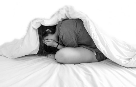 Black and white picture of girl with hands on face under white comforter