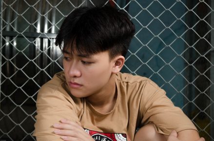 Asian boy looking sideways, distraught, in front of chain link fence