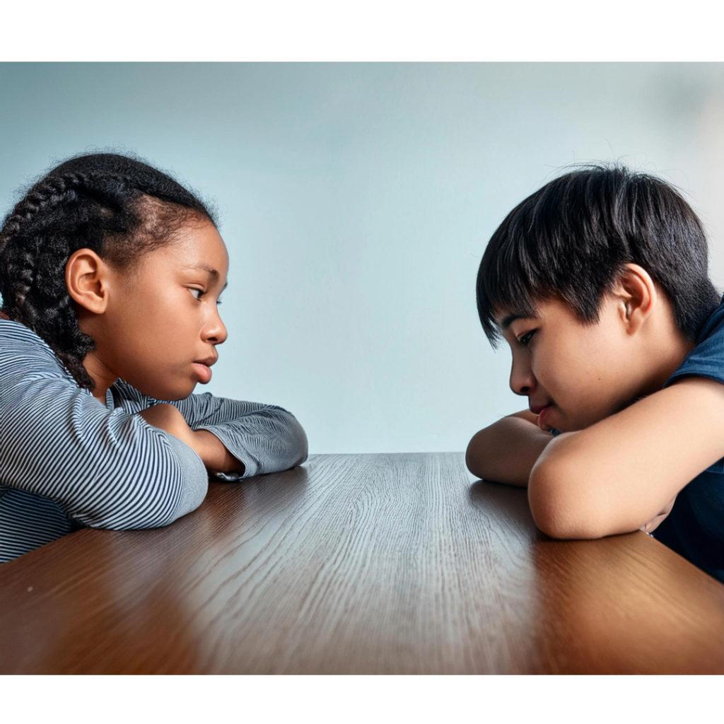 Two students sitting facing each other at table, unhappily